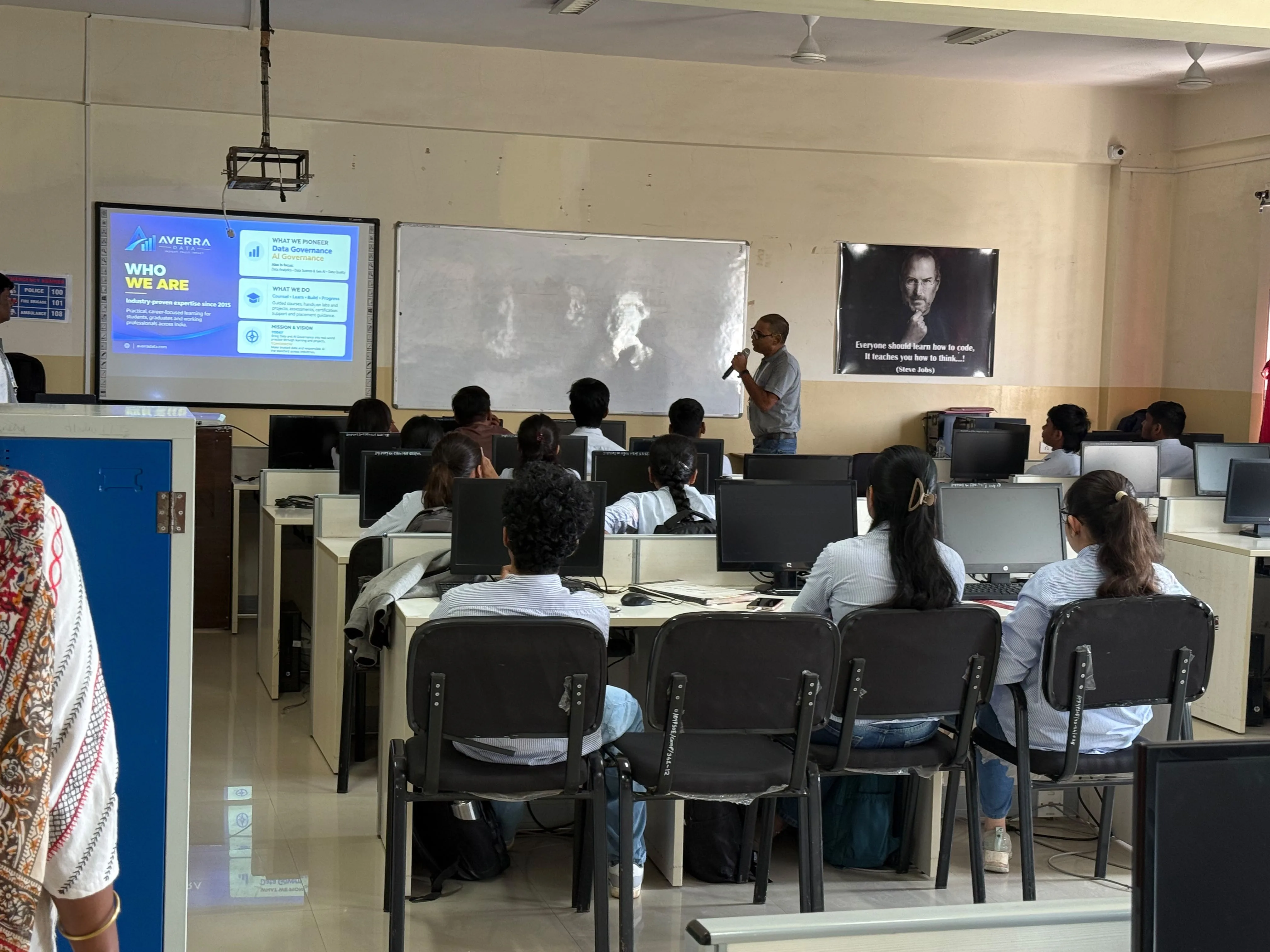 A presenter with a microphone introducing Averra Data to a classroom of students at their computers, with a Who We Are slide on the screen